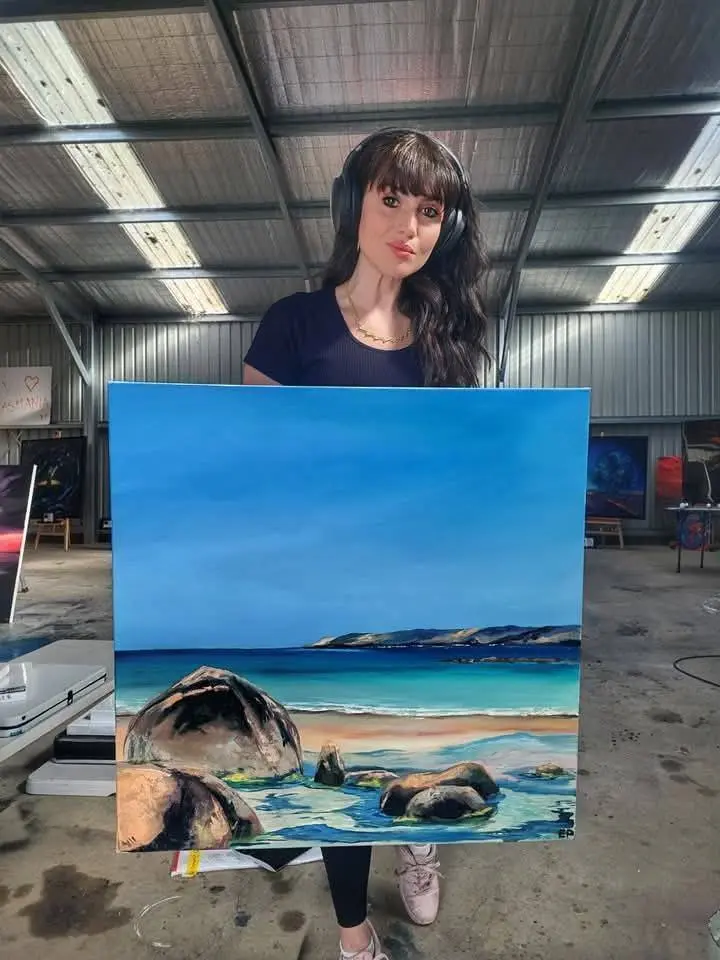 Artist holding large coastal landscape painting in studio