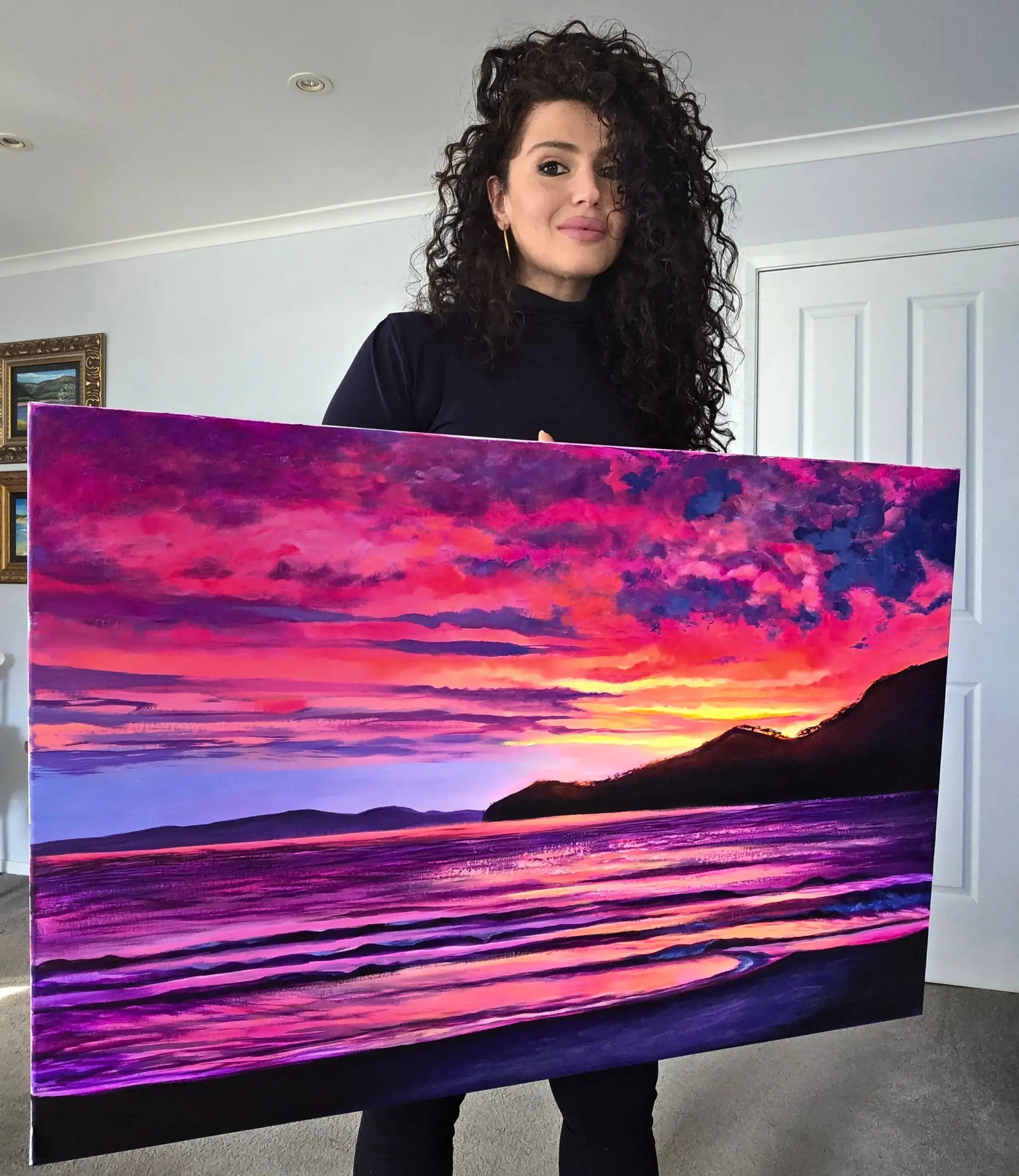 1000155707 Woman holding vibrant sunset seascape painting indoors